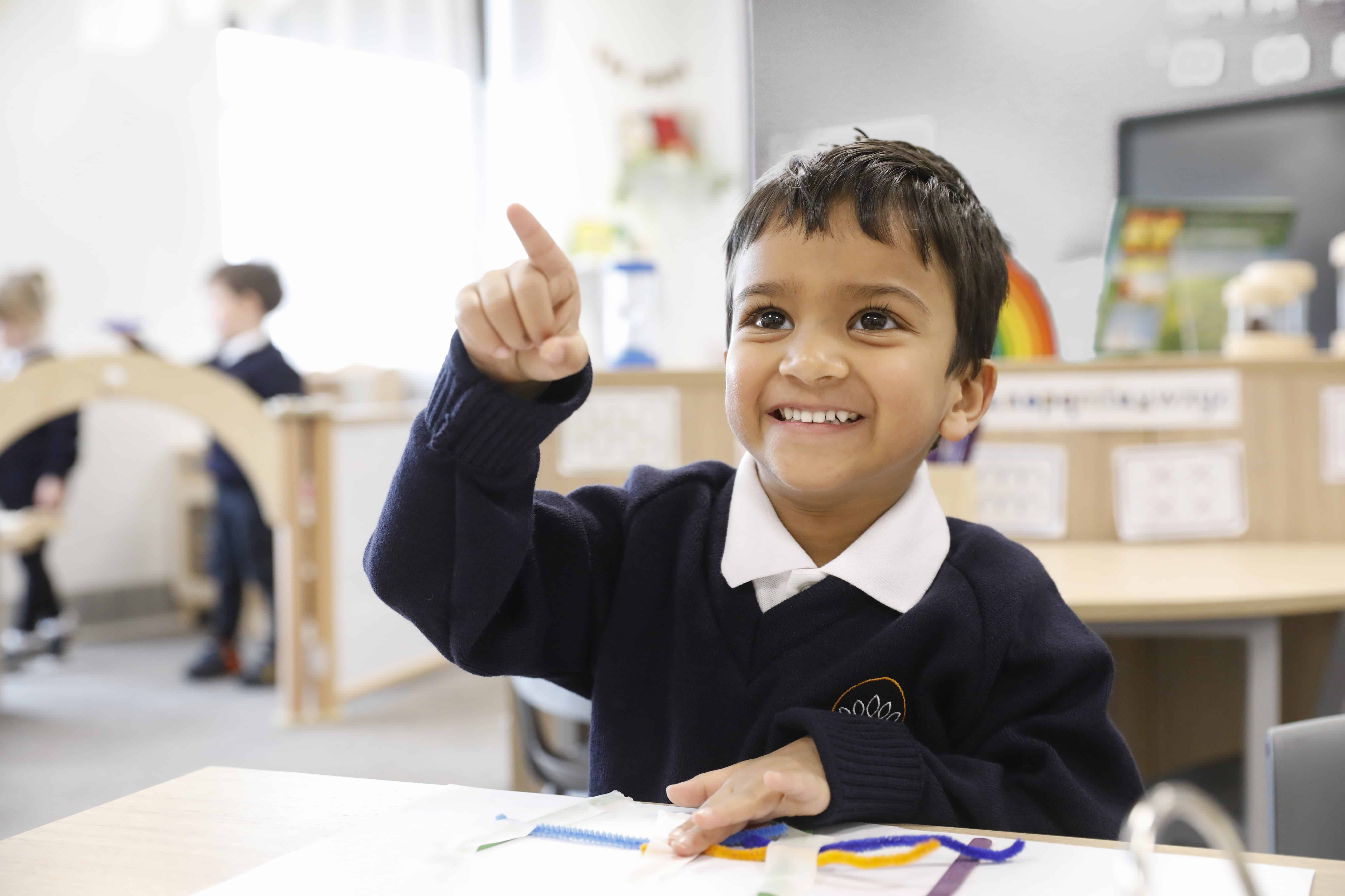 Image of Pre School pupil at Crown Street Primary School smiling with hand in the air whilst completing a craft activity.